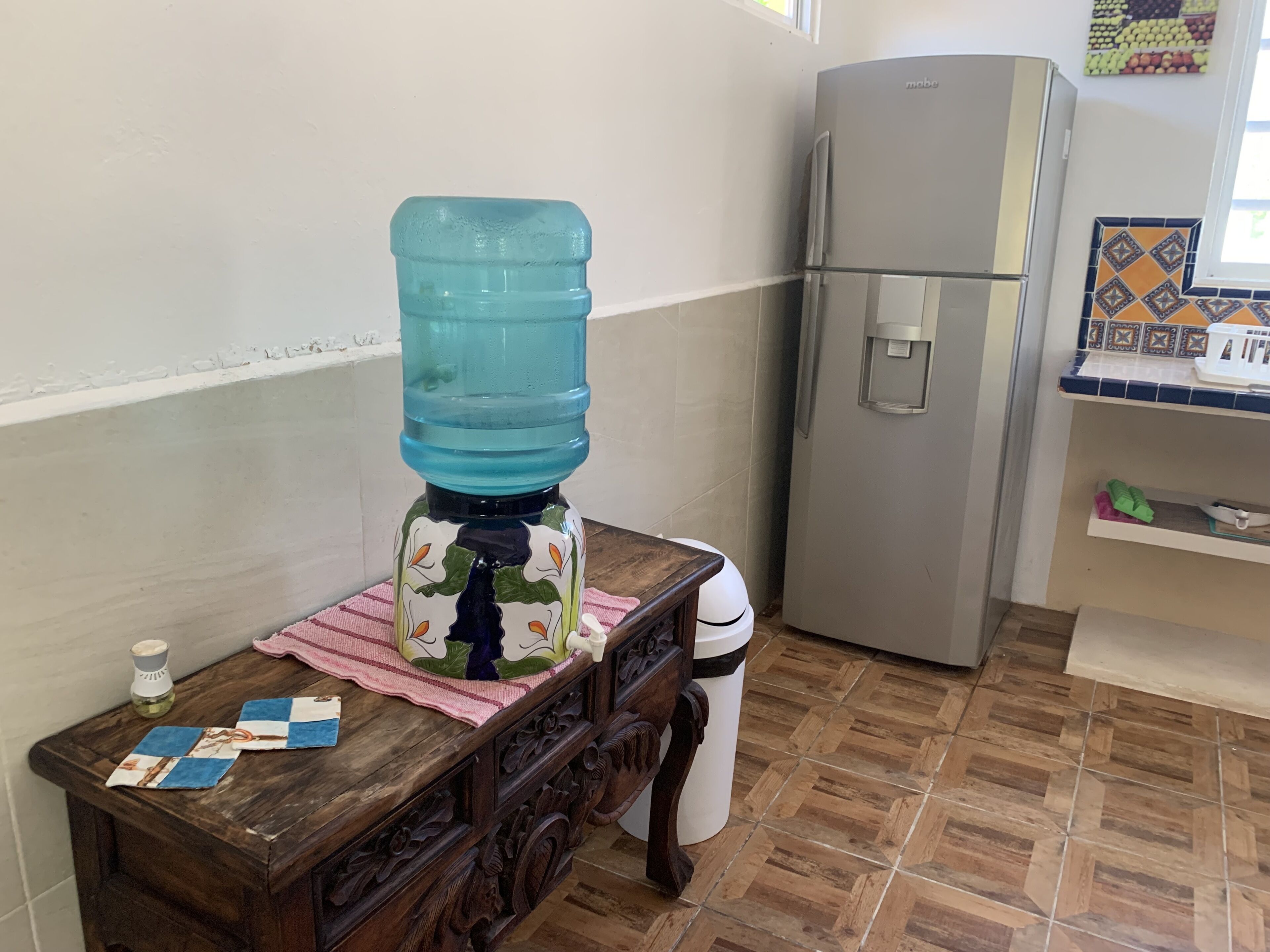 A kitchen with a water dispenser, a refrigerator, a wooden table with a cloth, and a tiled floor.