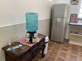 A kitchen with a water dispenser, a refrigerator, a wooden table with a cloth, and a tiled floor.