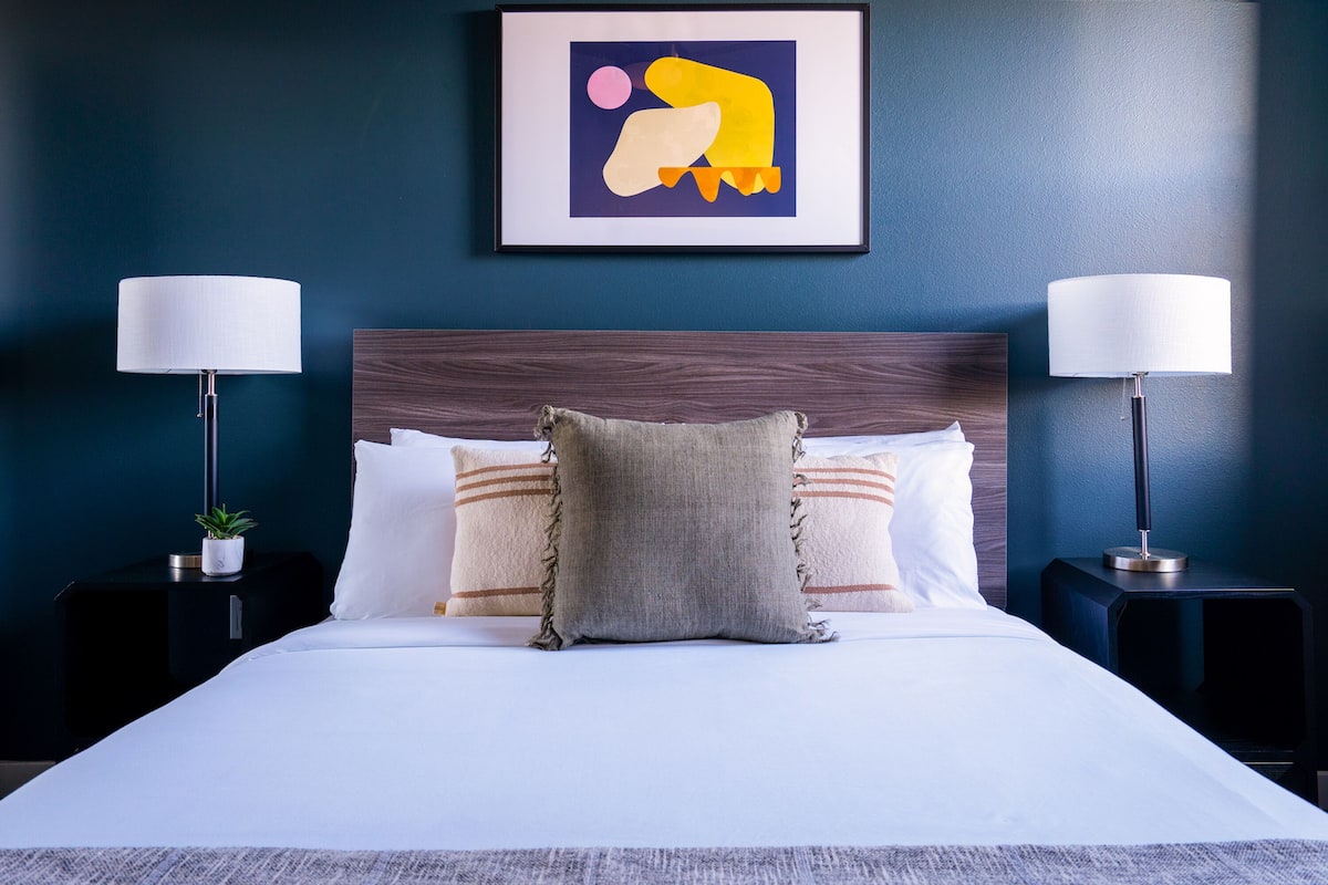 A neatly made bed with white linens and a gray throw pillow, flanked by two black nightstands with lamps, against a dark blue wall.