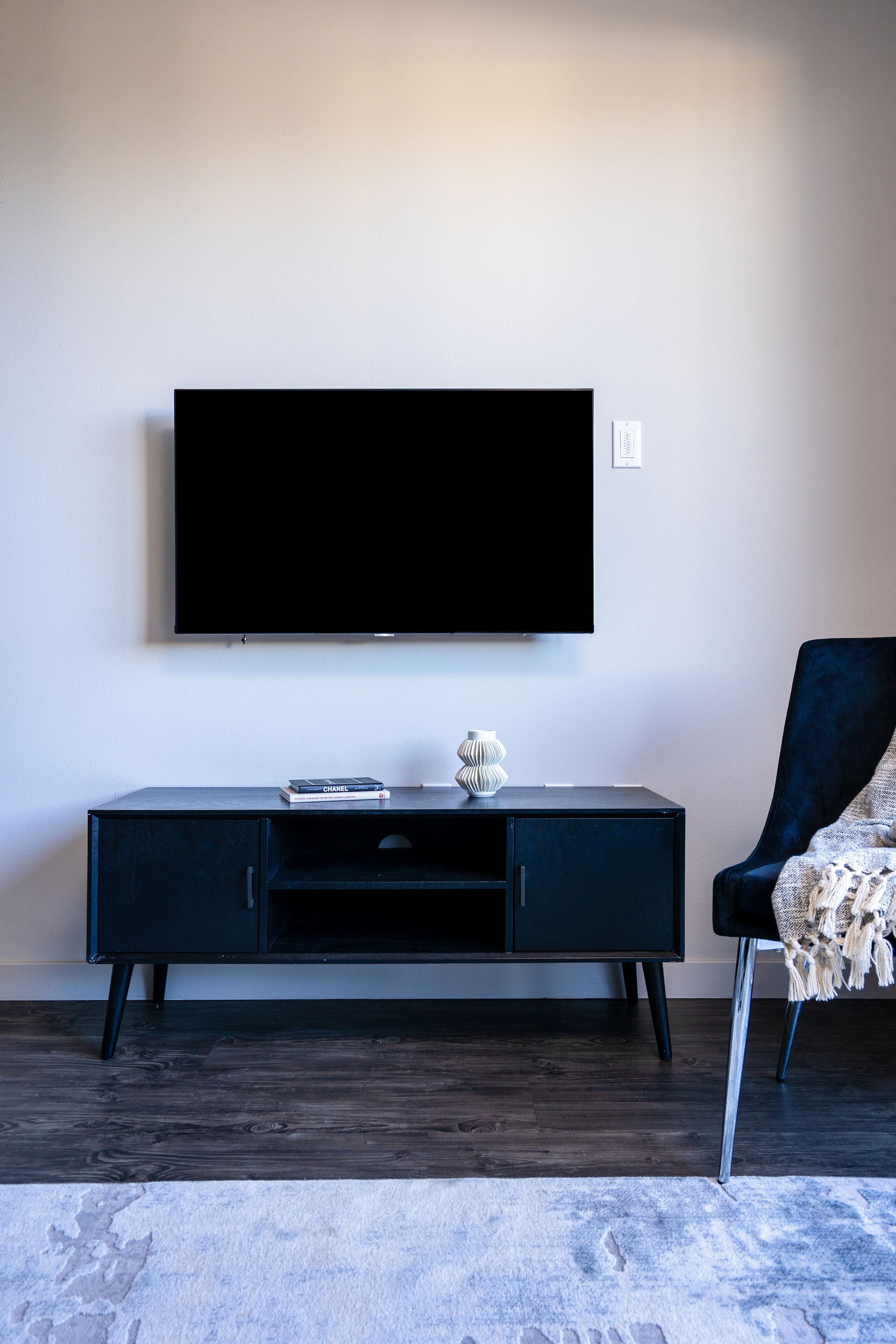 Un salon moderne avec un téléviseur à écran plat fixé au mur, un meuble en bois foncé en dessous, et un fauteuil bleu foncé agrémenté d’un plaid à motifs.
