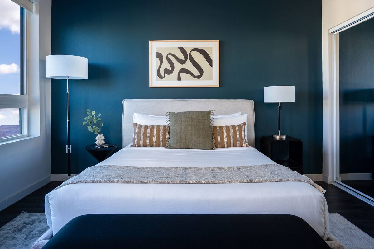 A modern bedroom with a large bed, two bedside tables, a floor lamp, and a wall-mounted abstract art piece.