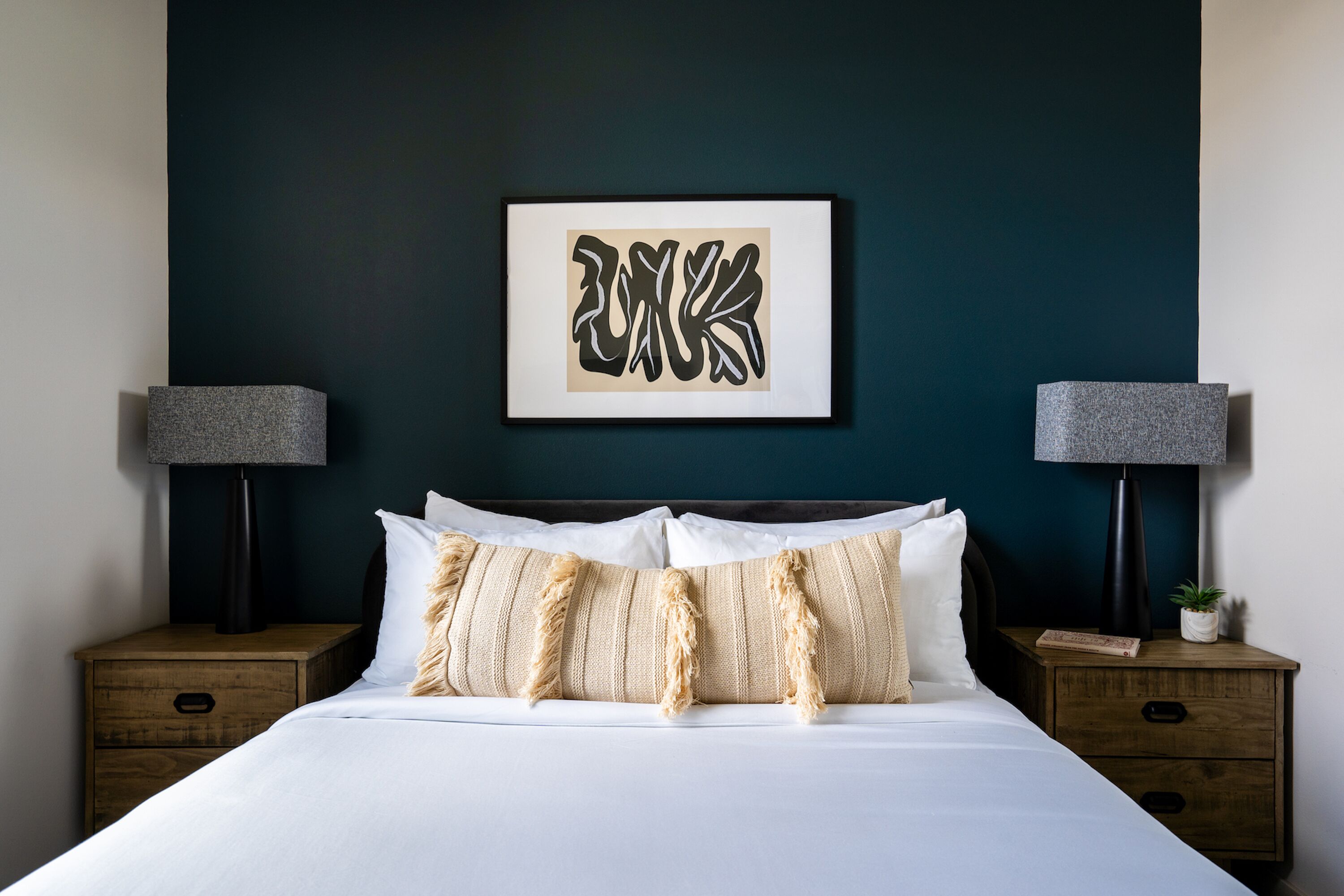 Una cama bien hecha con sábanas blancas y almohadas a rayas, con dos mesitas de noche a cada lado, con lámparas y una planta pequeña. Un cuadro enmarca una obra de arte sobre la cama.