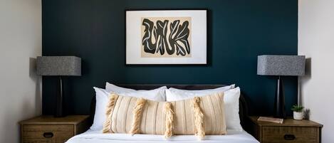 Una cama bien hecha con sábanas blancas y almohadas a rayas, con dos mesitas de noche a cada lado, con lámparas y una planta pequeña. Un cuadro enmarca una obra de arte sobre la cama.