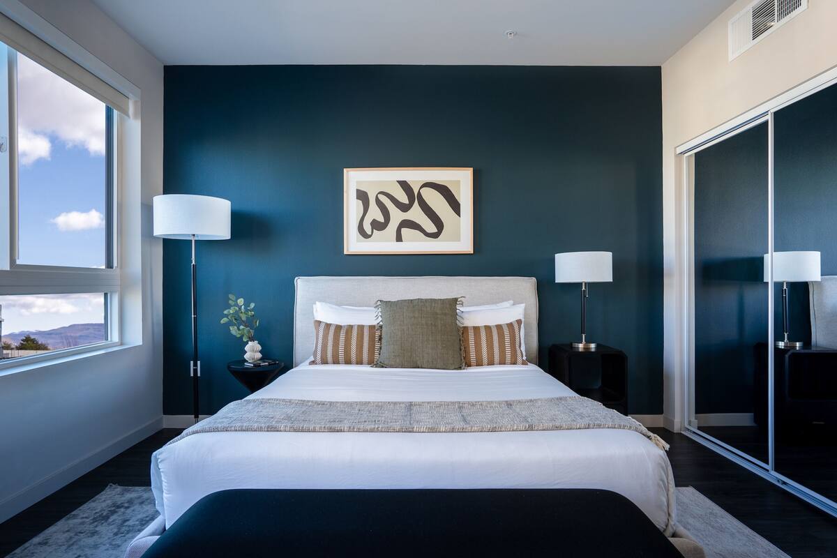 A modern bedroom with a large bed, two bedside lamps, a framed abstract artwork, and a view of the outdoors through a window.