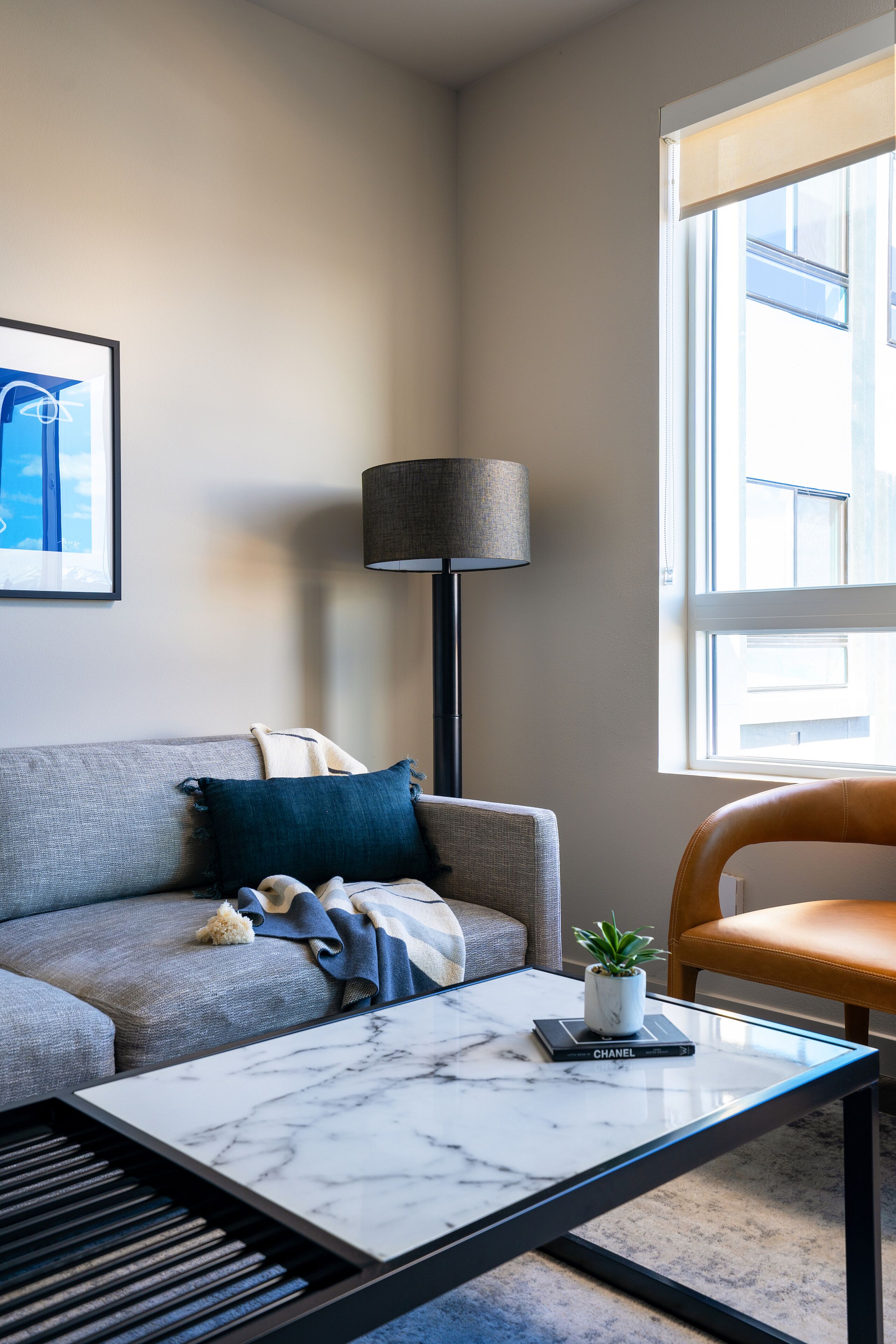 Un salon moderne avec un canapé gris, un lampadaire noir, un fauteuil en cuir, une table basse en marbre et une petite plante en pot.