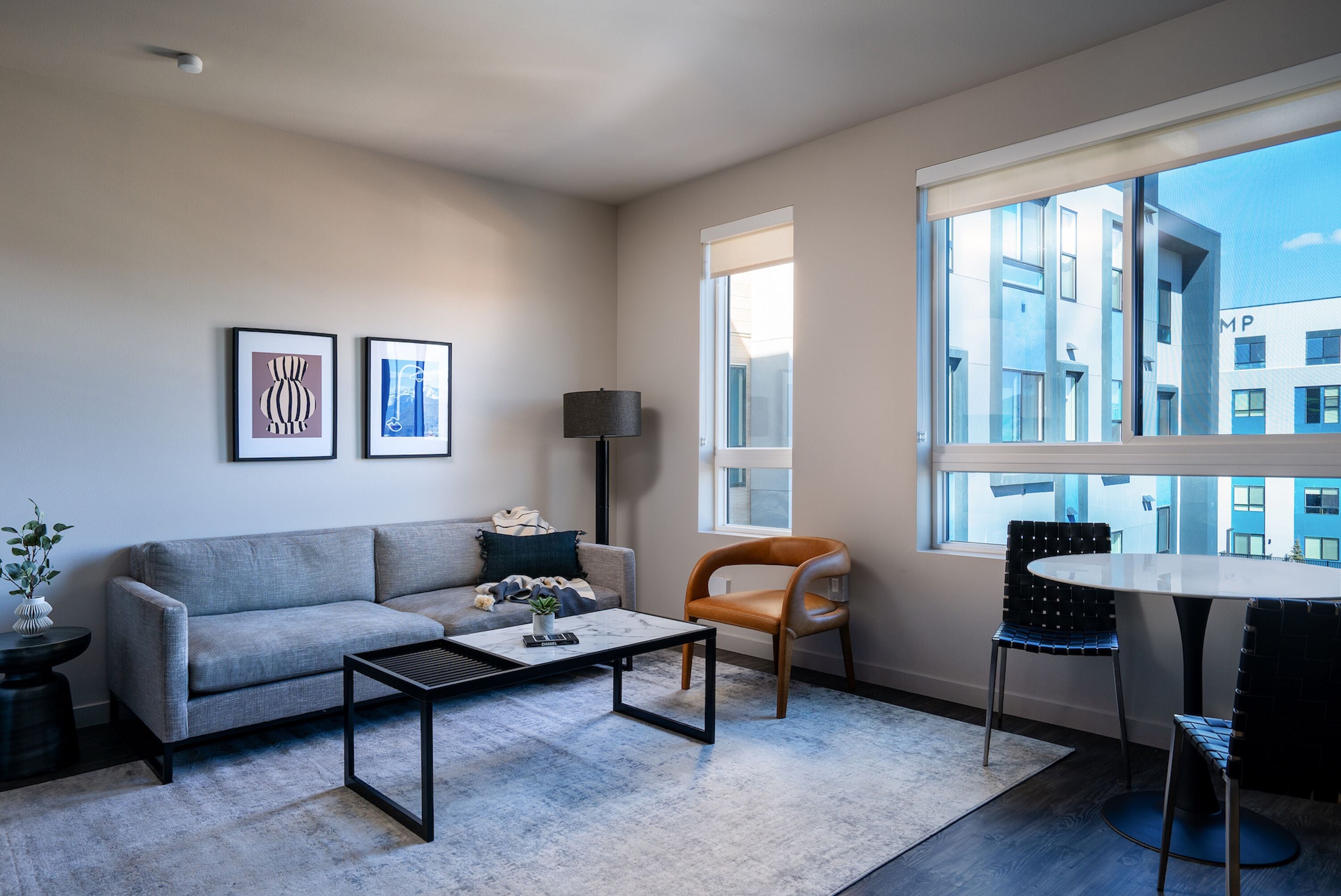 A modern living room with a grey sofa, a coffee table, a side table with a plant, and a lamp. There is a window with a view of buildings and a dining area with a table and chairs.