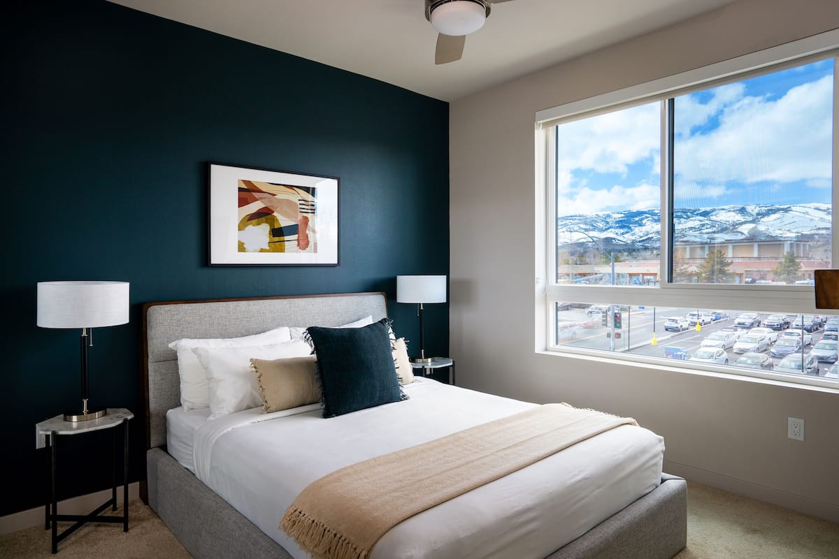 A modern bedroom with a large bed, two bedside lamps, a ceiling fan, a window with a view of a parking lot and mountains.