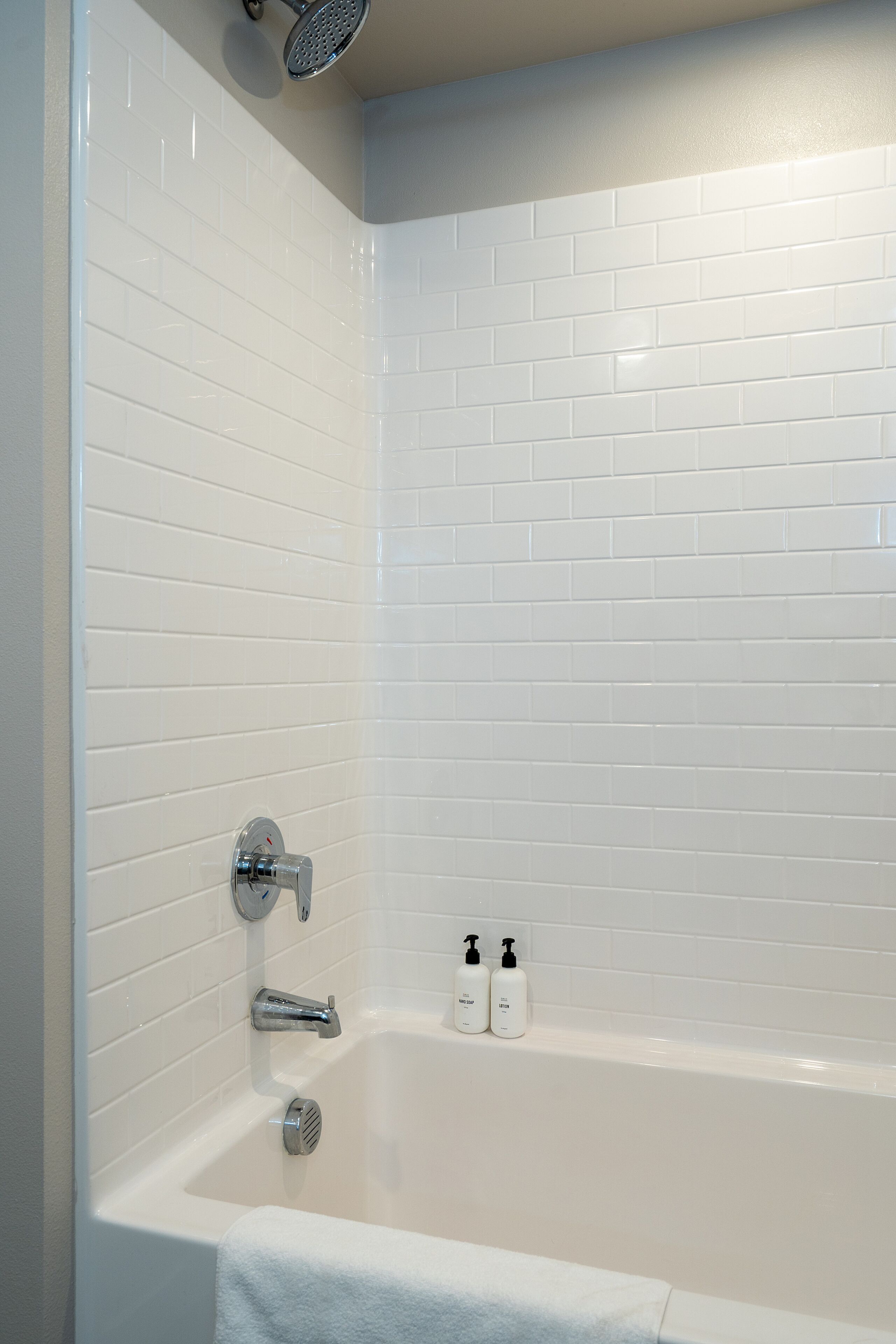 Un baño con paredes de azulejos blancos, una bañera, un cabezal de ducha y dos botellas de champú.