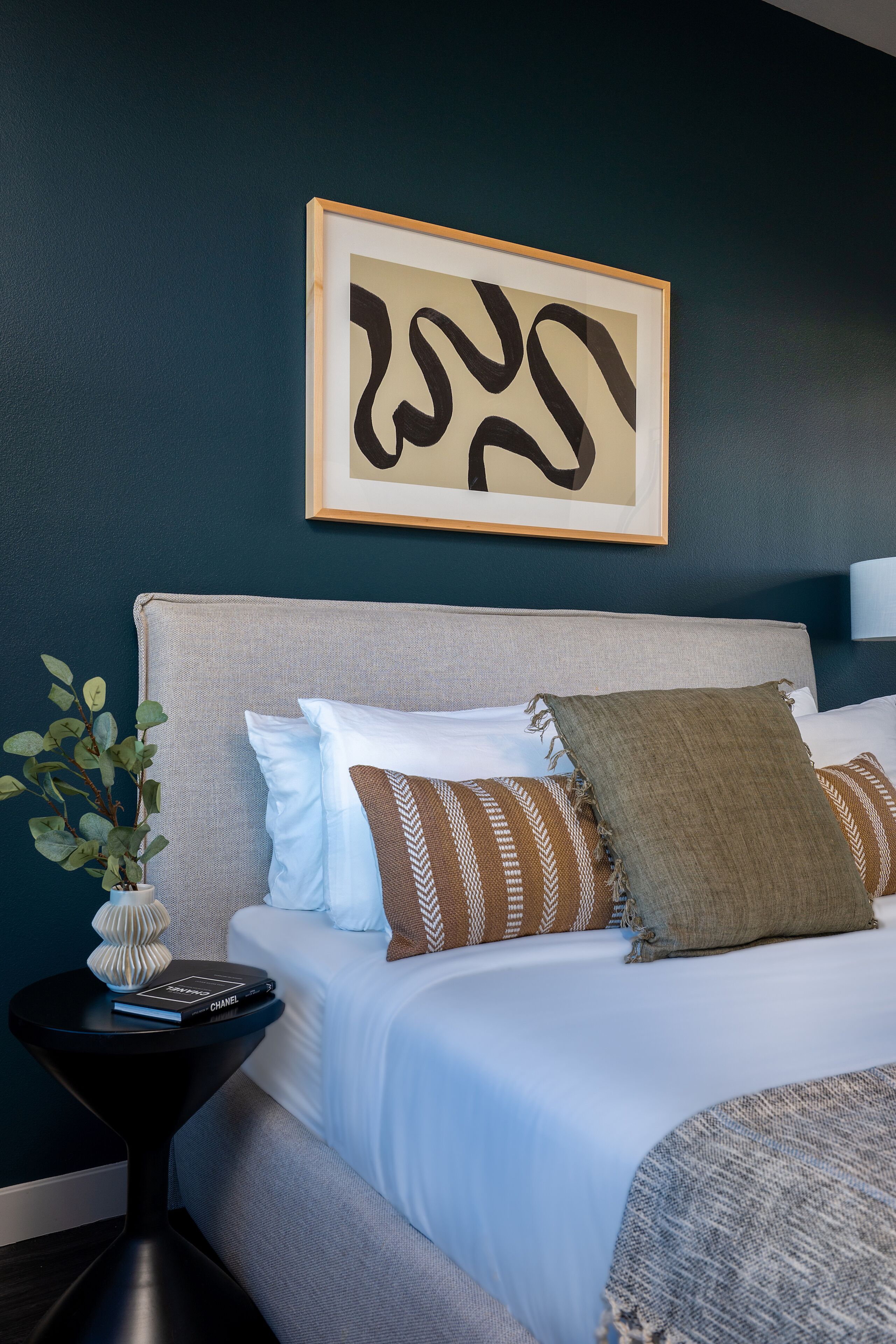 A neatly made bed with a grey headboard, a framed abstract artwork above it, and a small side table with a plant and a book.
