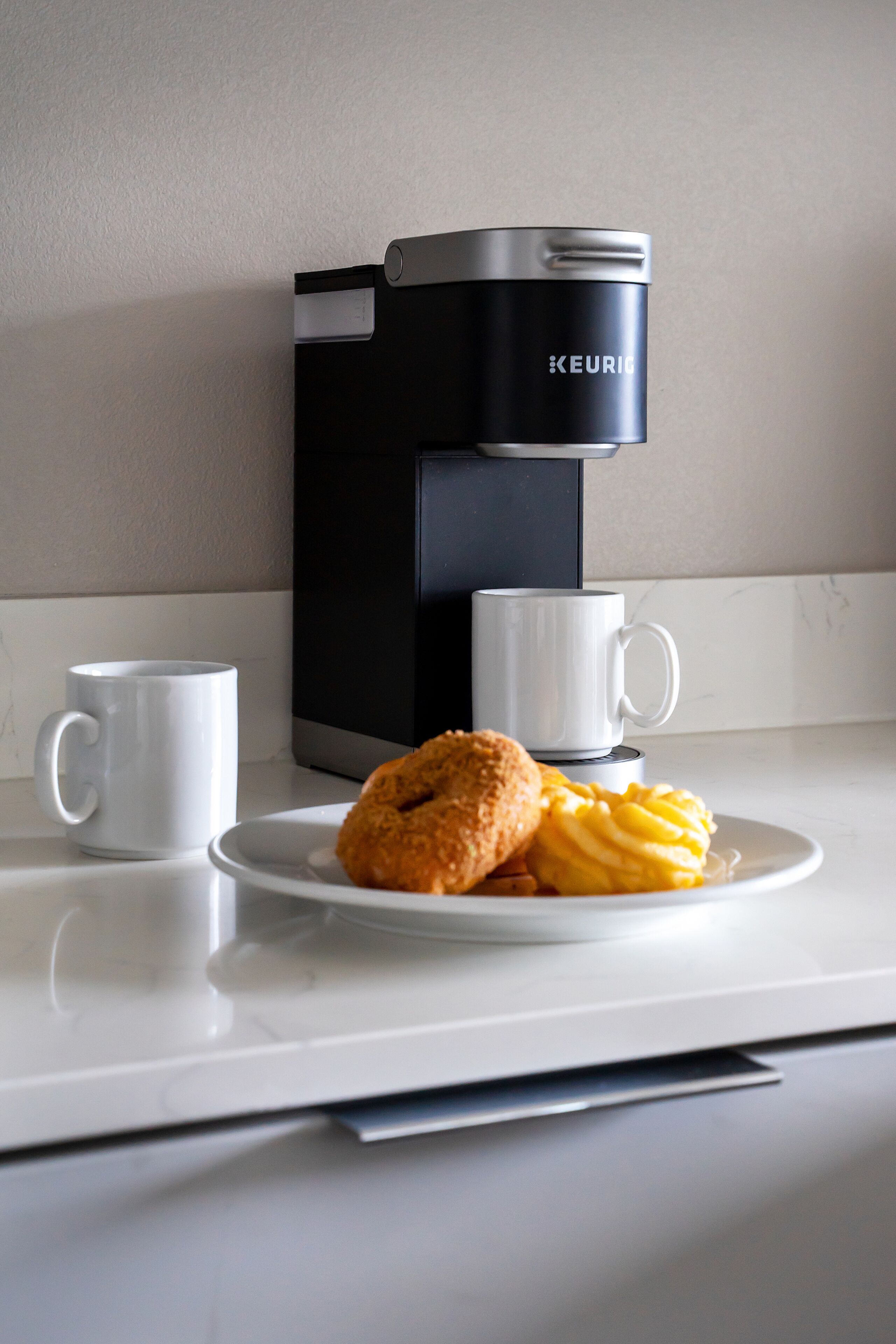A modern coffee maker on a countertop with two white mugs and a plate with pastries.