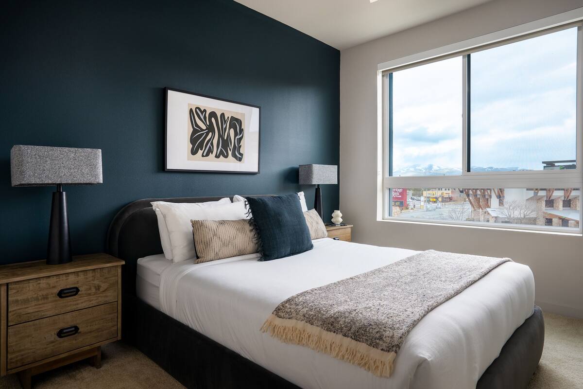 A modern bedroom with a large bed, two bedside tables with lamps, a framed artwork on the wall, and a window offering a city view.
