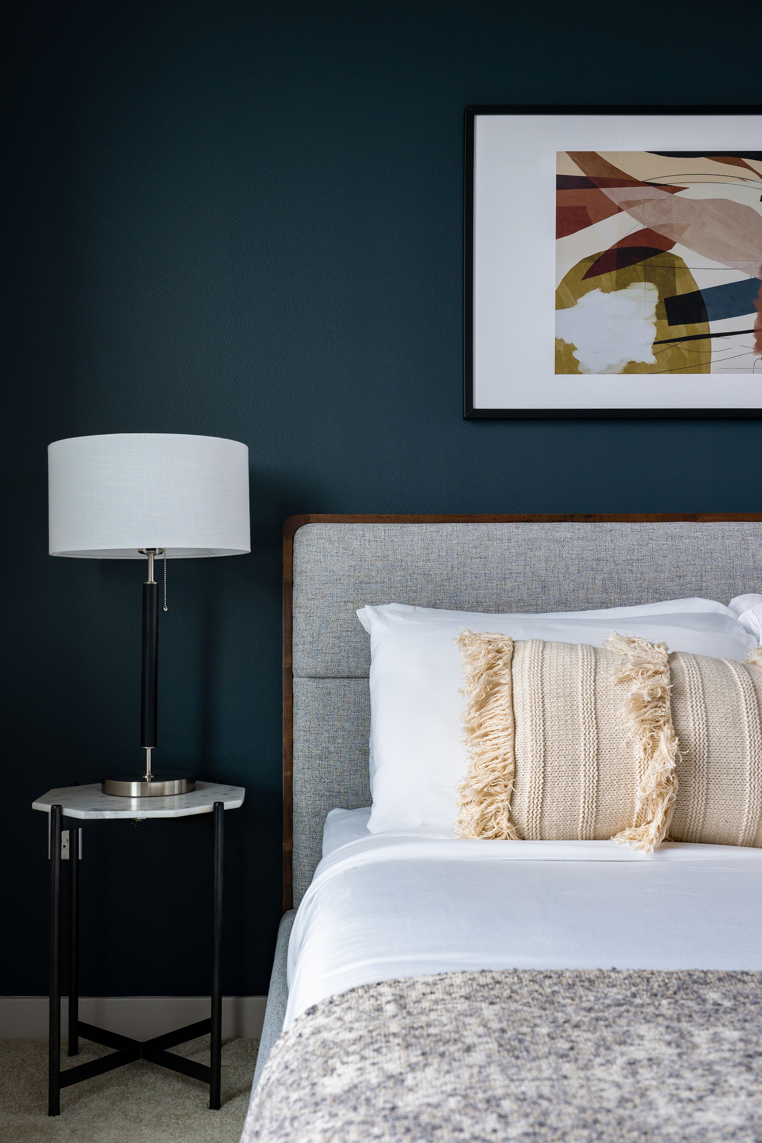 Een netjes opgemaakt bed met witte lakens en een beige plaid, een moderne vloerlamp en een lijst met een abstract schilderij aan de muur.