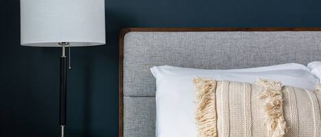 A neatly made bed with white linens and a beige throw blanket, a modern floor lamp, and a framed abstract painting on the wall.