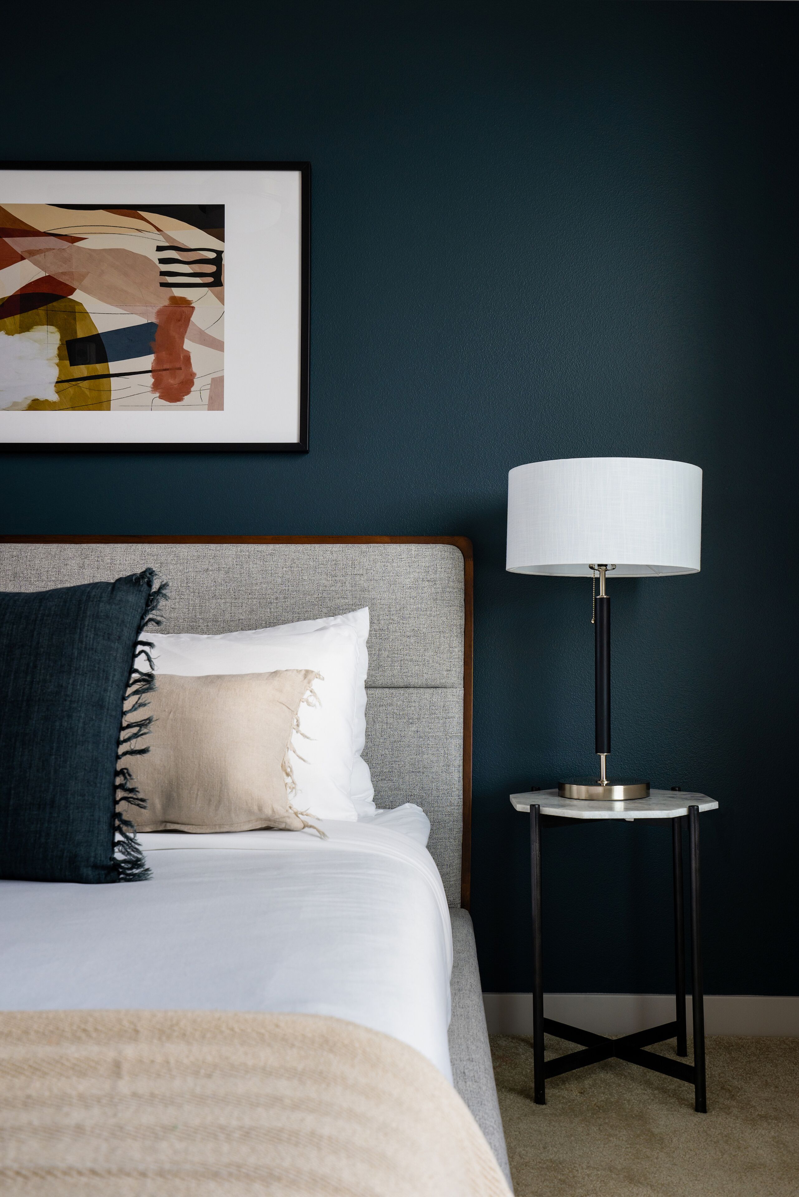 A neatly made bed with a grey headboard, a white pillow, and a blue throw blanket. A modern floor lamp with a white shade is placed beside the bed.
