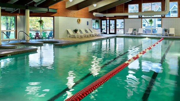 An indoor swimming pool with clear lane dividers, lounge chairs, and a view of the outside through large windows.
