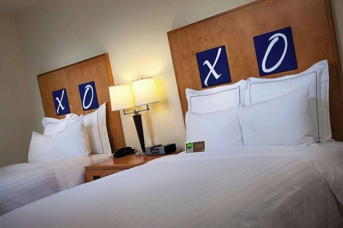A hotel room with two beds, each with a headboard displaying the letters 'X' and 'O'. There is a bedside table with a lamp, a phone, and a small sign.