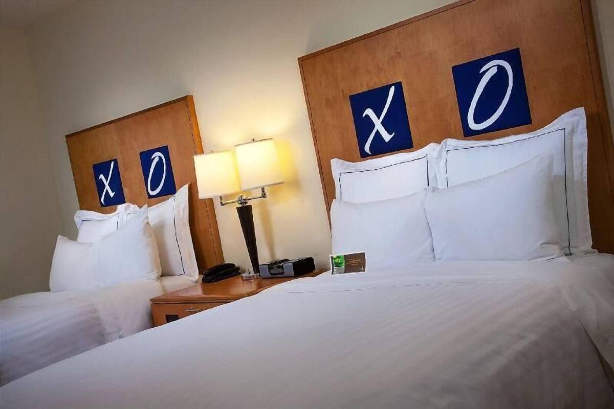 A hotel room with two beds, each with a headboard featuring a large 'X' and 'O' sign. There is a bedside table with a lamp and a phone.