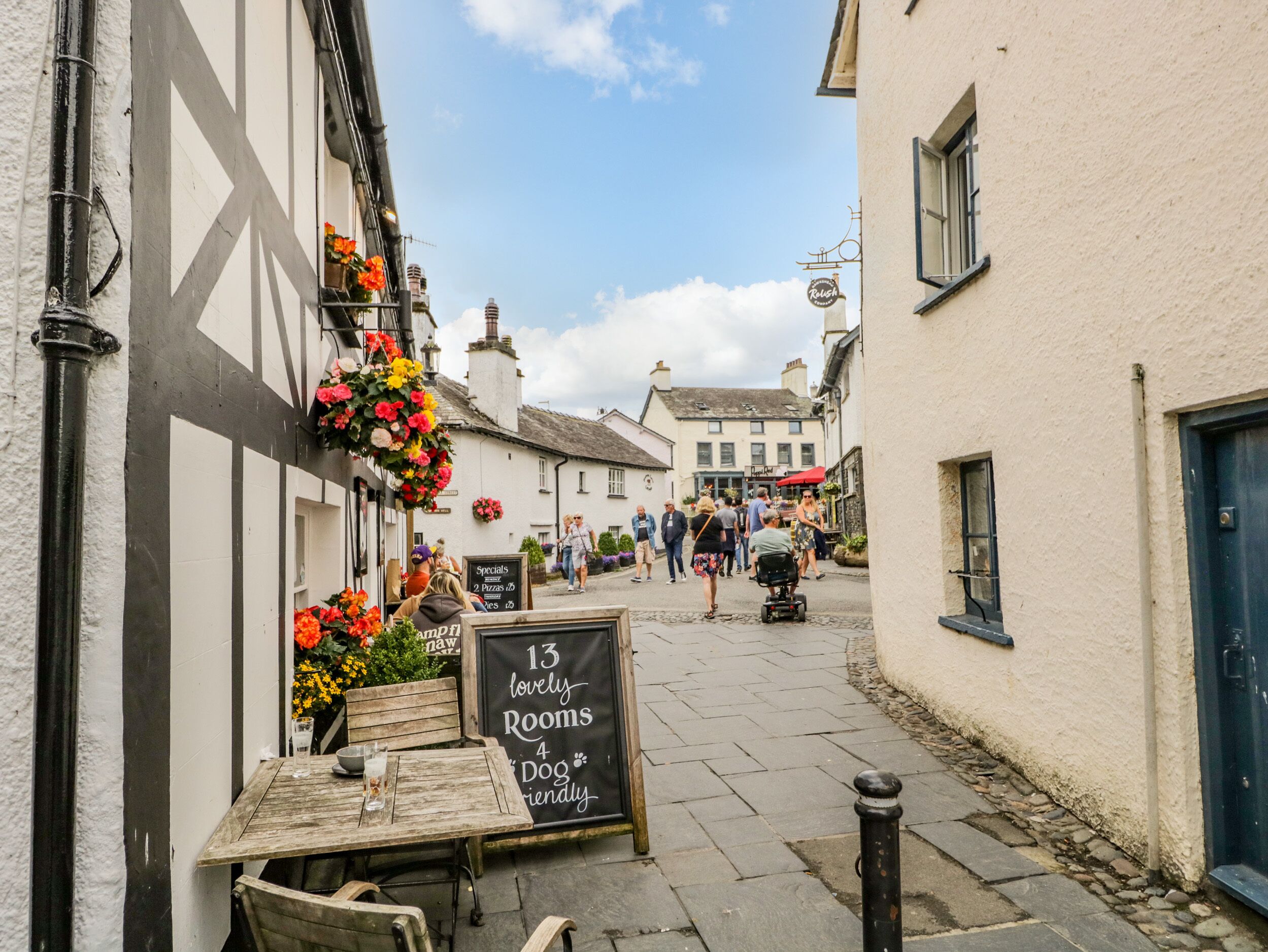 A narrow street with a pub on the left, a sign for '13 lovely rooms' and '4 dog friendly', and people walking in the distance.