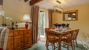 A dining area with a wooden table set for four, flanked by wooden chairs. A wooden cabinet with drawers is to the left, and a lamp with a red base is on the table. A mirror hangs on the wall above the table.