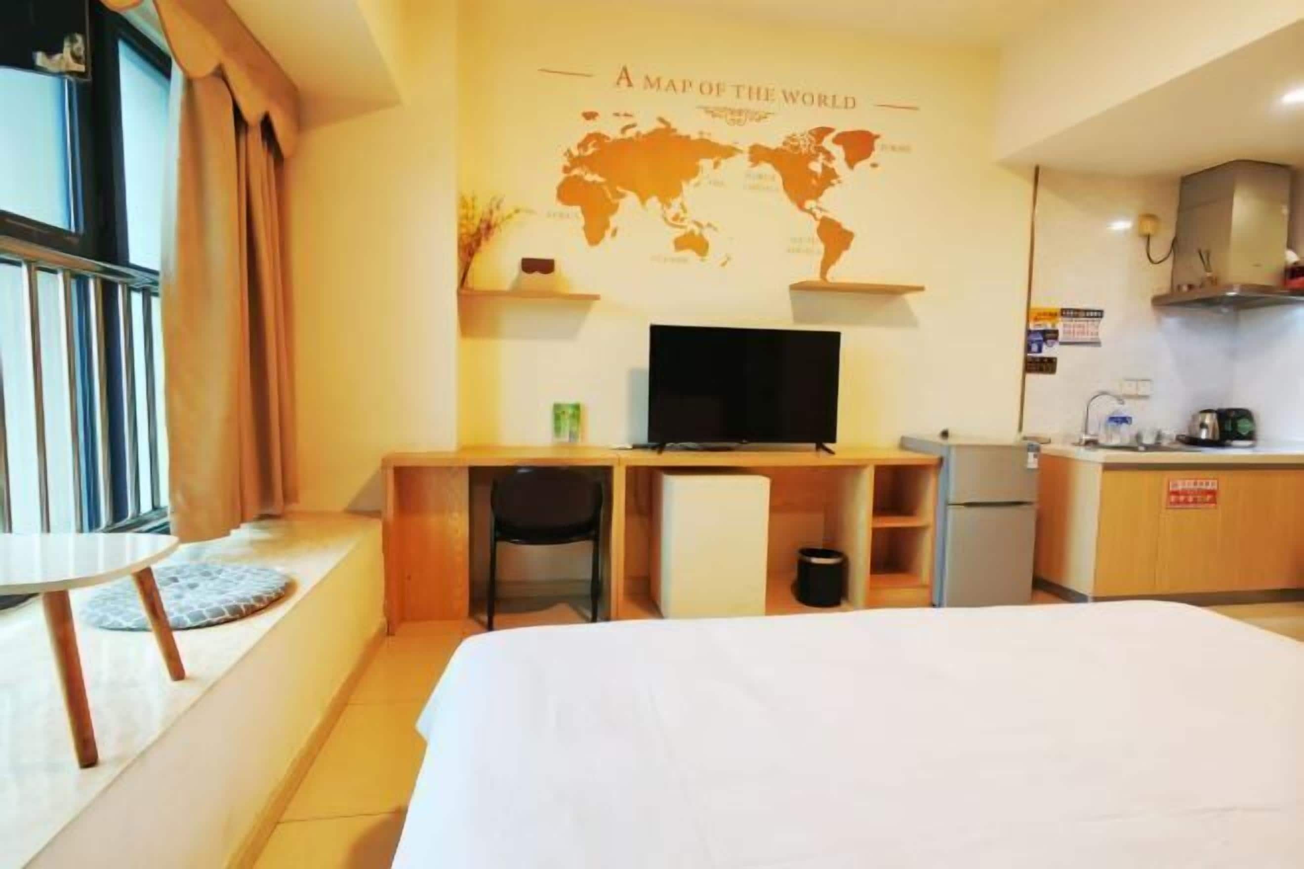 Habitación de hotel con cama, escritorio con computadora, kitchenette pequeña y un mapa mundial en la pared.