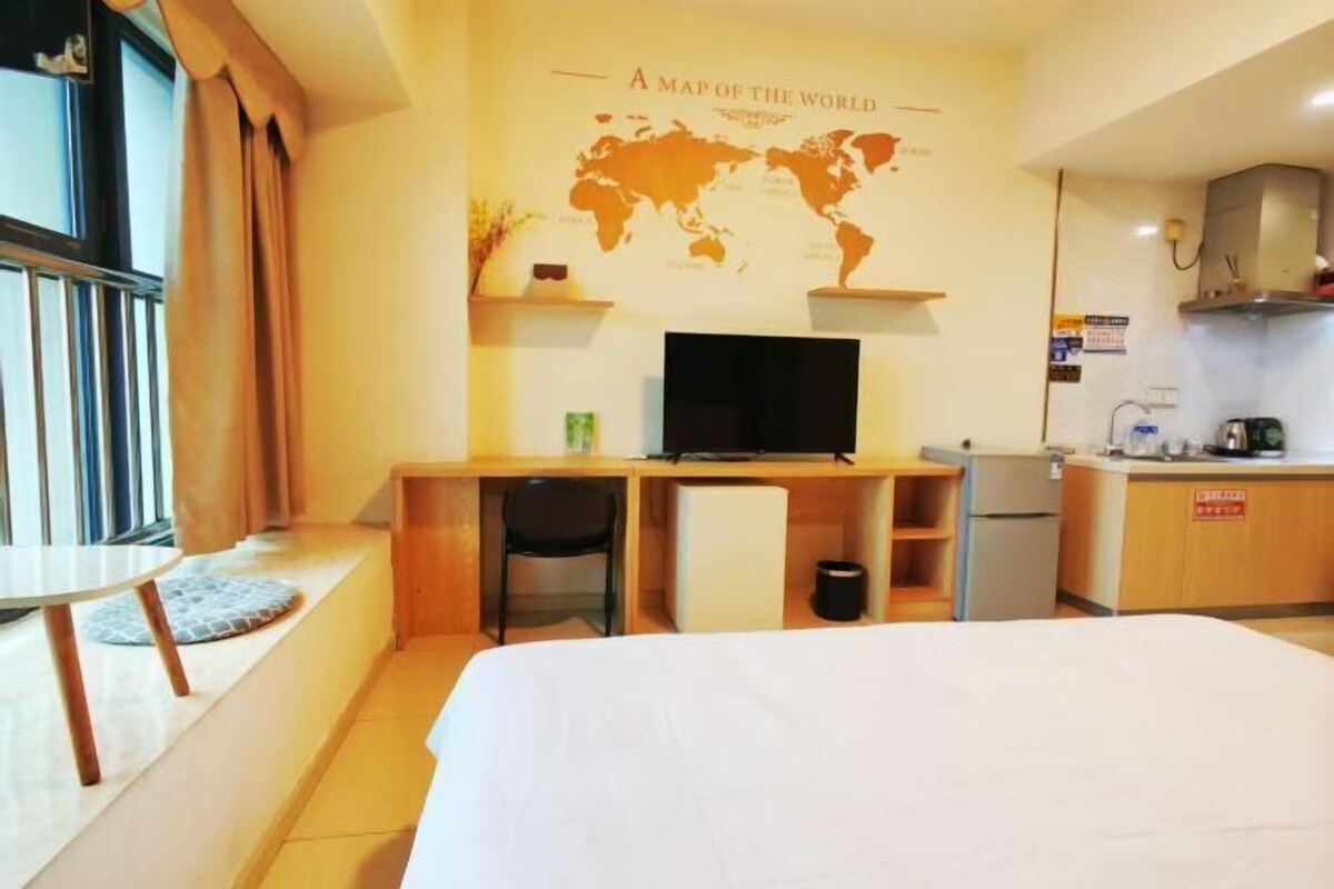 A hotel room with a bed, a desk with a computer, a small kitchenette, and a world map on the wall.