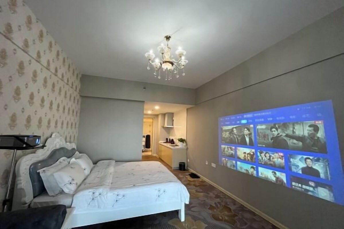 A hotel room with a bed, a chandelier, and a wall-mounted screen displaying movie scenes.