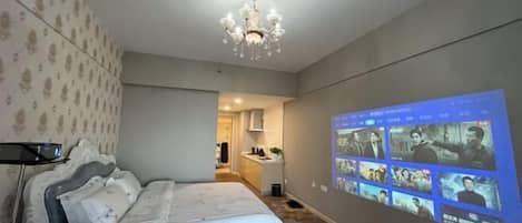 A hotel room with a bed, a chandelier, and a wall-mounted screen displaying movie scenes.