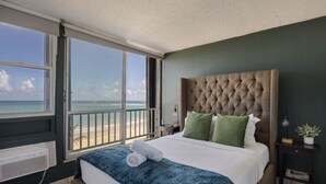 A bedroom with a large window offering a view of the beach, a tufted headboard, and a neatly made bed with green pillows.