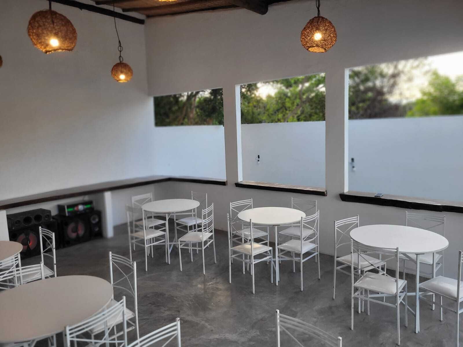 A room with white tables and chairs arranged in a circular pattern, large windows with a view of greenery, and hanging pendant lights.
