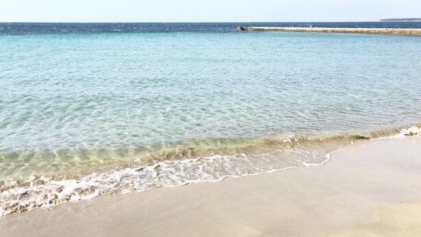 Ein Strand mit klarem Wasser, sandigem Ufer und einem in der Ferne liegenden Wellenbrecher.