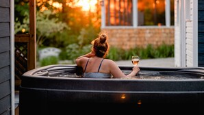Une personne se détend dans un bain à remous, un verre à la main, entourée de verdure et avec une maison en arrière-plan.