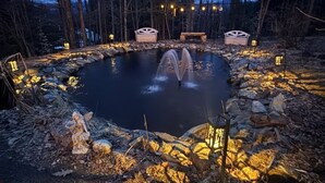 Un bassin circulaire avec une fontaine, entouré de rochers et illuminé par des guirlandes lumineuses.