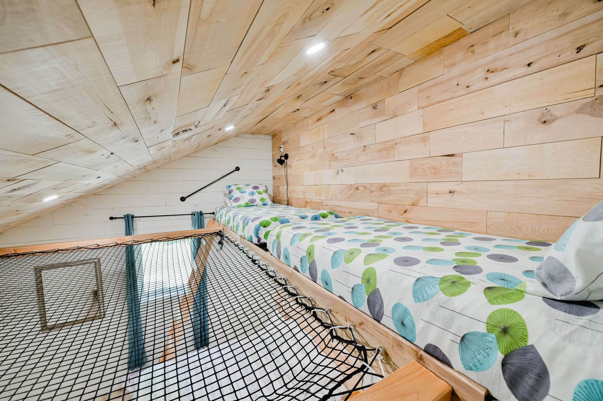 A room with a sloped ceiling, wooden walls, a bed with a patterned bedspread, and a netted window blind.