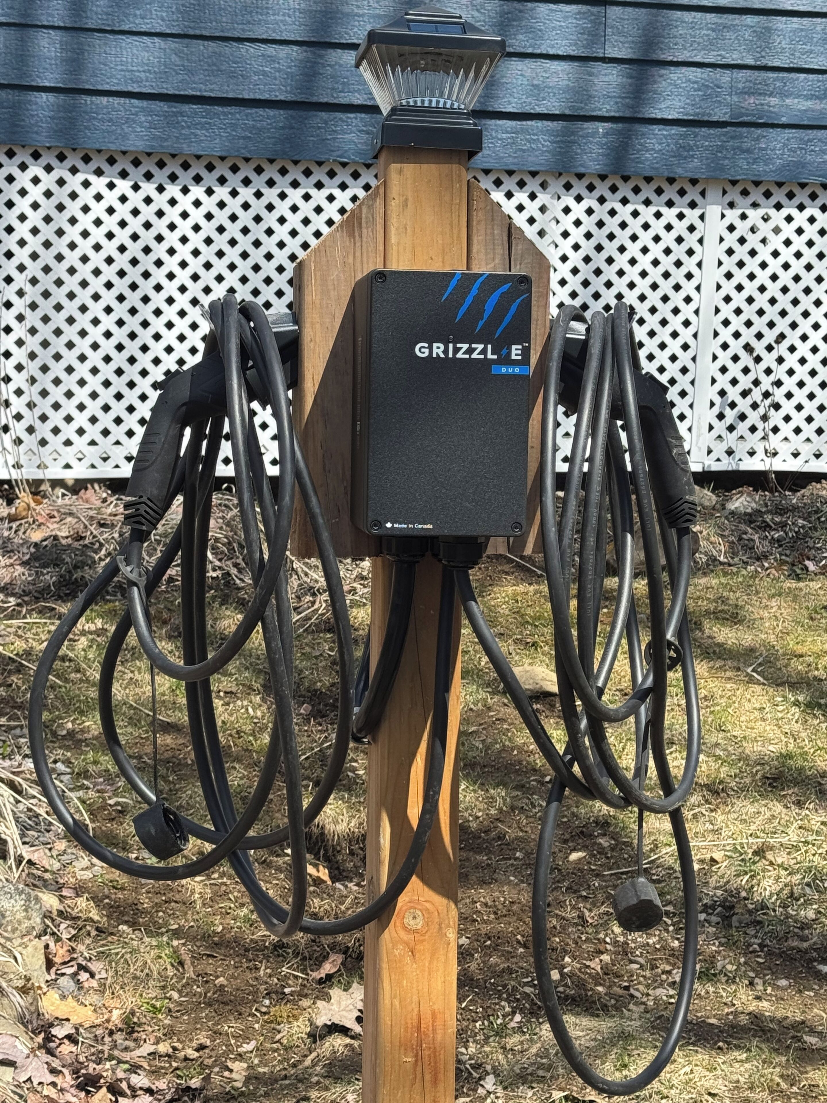 A wooden outdoor electrical box with multiple cables connected to it, labeled "GRIZZLE Duo," against a white lattice fence.