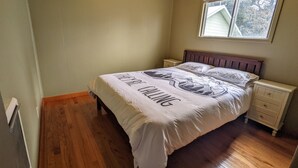 Une chambre avec un plancher en bois, un lit recouvert d’une courtepointe blanche, une table de chevet et une fenêtre avec des stores.