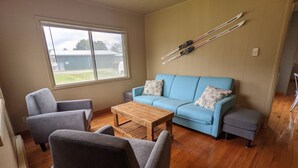 Un salon avec un canapé bleu, deux fauteuils, une table basse en bois et une fenêtre donnant sur une maison et des arbres.