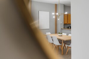 Un comedor moderno con una mesa de madera y sillas blancas, una lámpara colgante y una cocina al fondo.