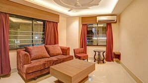 Ein modernes Wohnzimmer mit einer Couch, einem Couchtisch und einem Sessel. Große Fenster mit Vorhängen, eine Klimaanlage und ein Deckenventilator sind vorhanden.