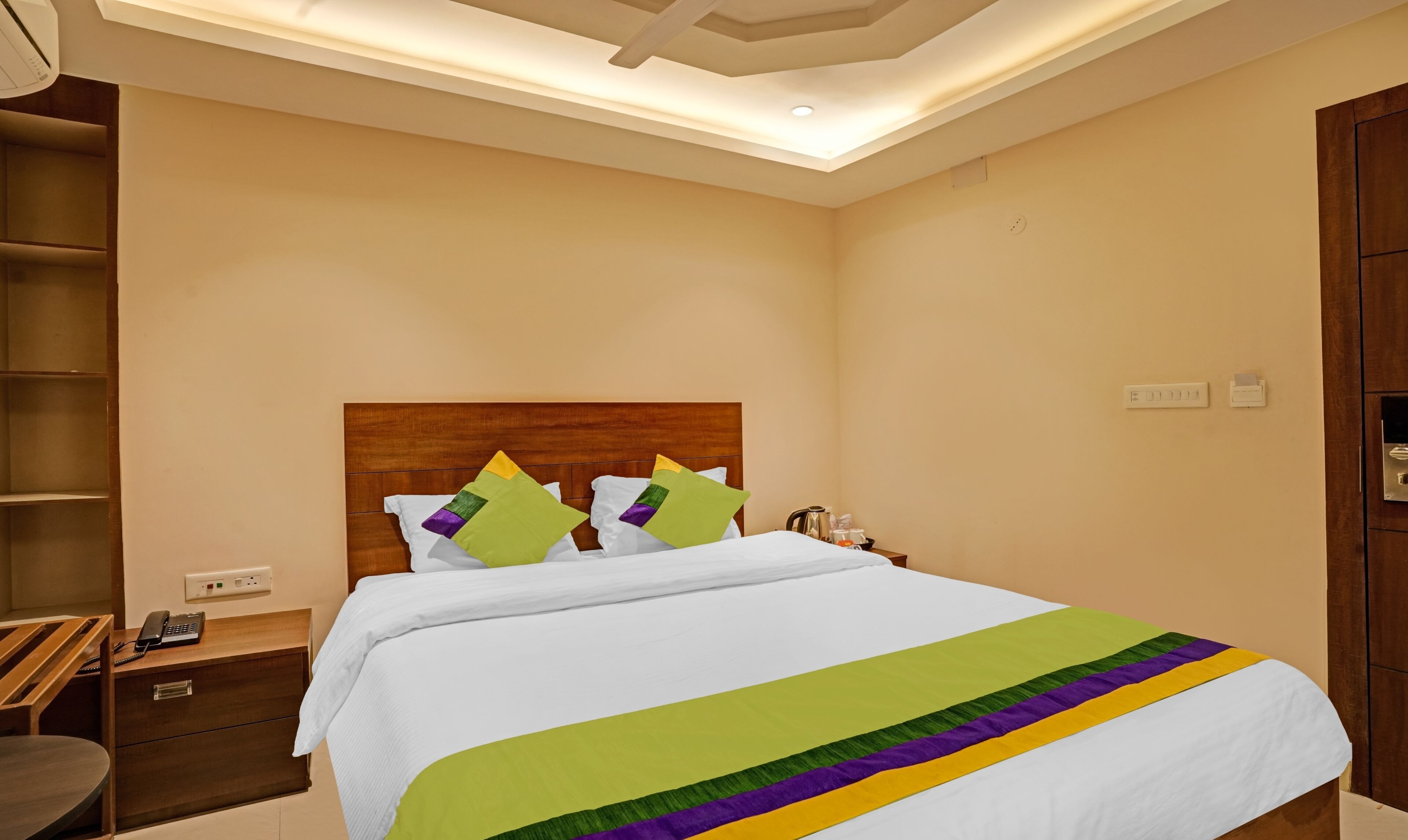 Habitación de hotel con cabecera de madera, cama con ropa de cama blanca y una manta colorida, una mesita de noche con un teléfono y una mesita auxiliar de madera.