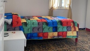 A small room with a bed covered in a colorful quilt, a white desk, and a window with curtains. - Apartment (Imperia)