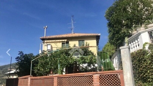 A two-story building with a red-tiled roof, a visible antenna, and a green fence with lattice panels. - Apartment (Imperia)