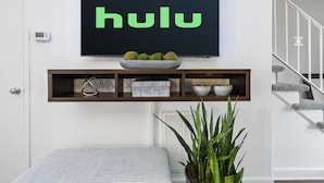 A modern living room with a large flat-screen TV mounted on the wall displaying the Hulu logo. Below the TV, there is a wooden shelf with decorative items and a potted plant. - Cozy Condo * Office* Near Luxury Mall & Uptown (Charlotte)