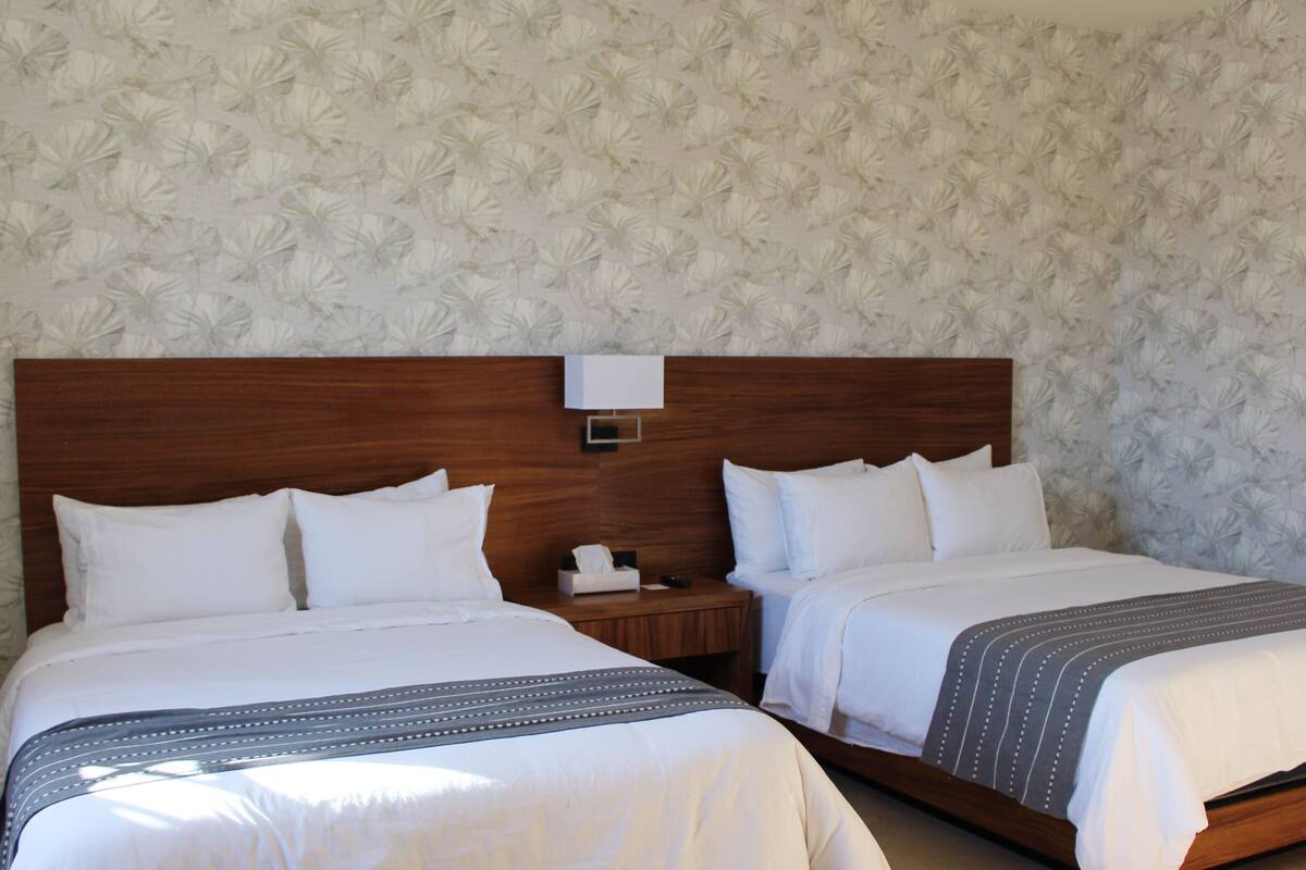 Une chambre d’hôtel avec deux lits, des têtes de lit en bois, du linge de lit blanc et un papier peint à motifs floraux.