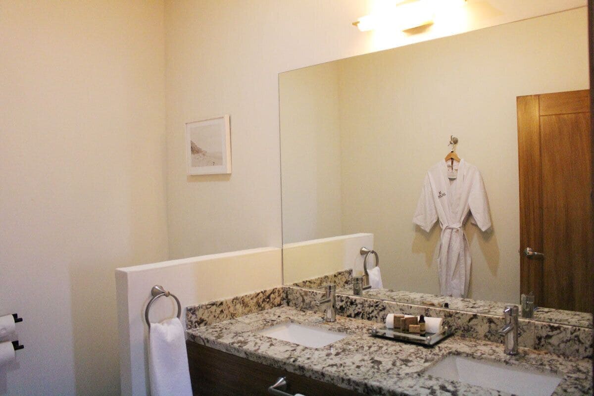 Une salle de bain avec un comptoir en granit, deux lavabos, un grand miroir, un porte-serviettes et une porte en bois.