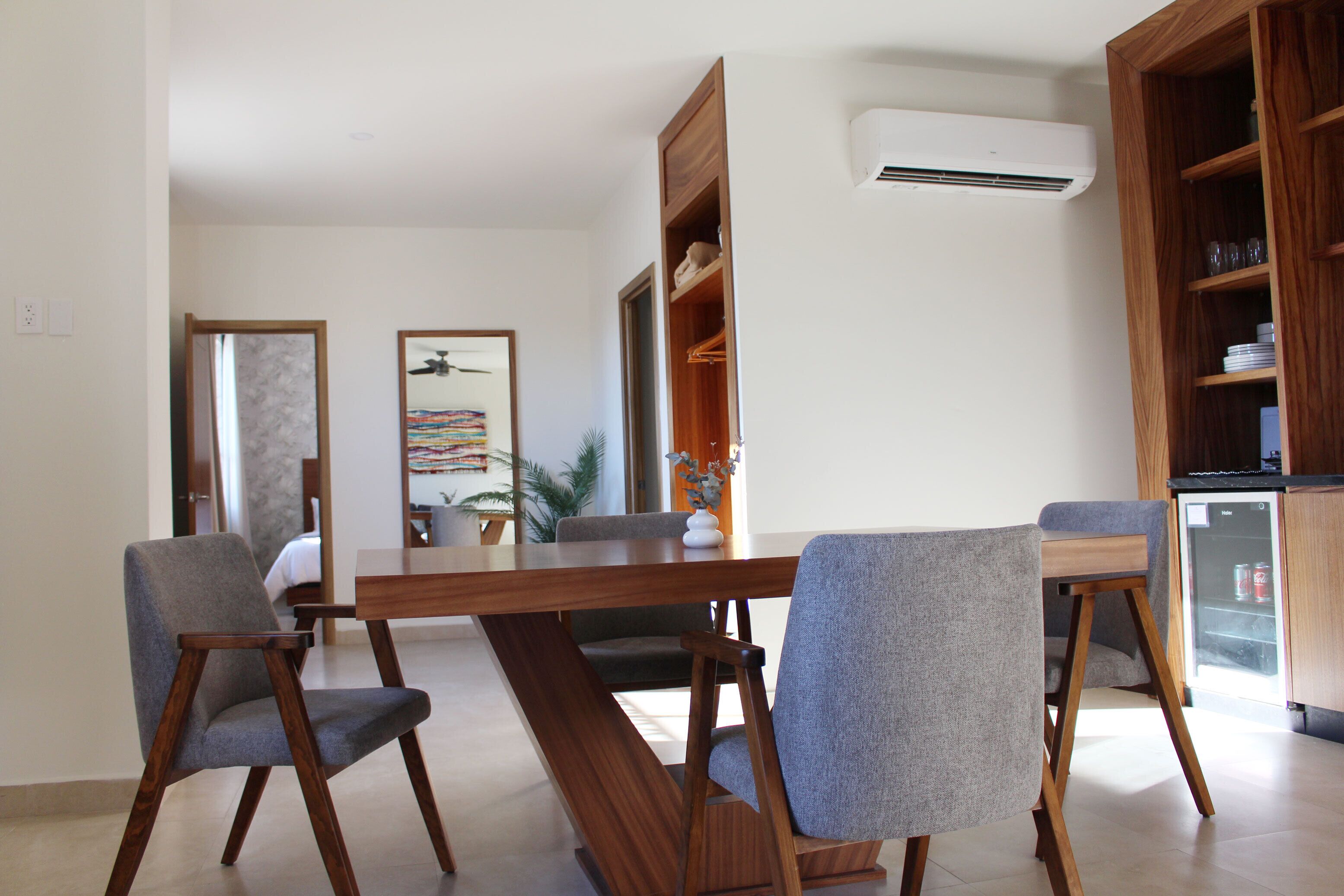 Un área de comedor moderna con una mesa de madera y sillas grises tapizadas. Se aprecia una estantería empotrada y un refrigerador pequeño al fondo.