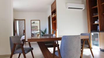 Un área de comedor moderna con una mesa de madera y sillas grises tapizadas. Se aprecia una estantería empotrada y un refrigerador pequeño al fondo.