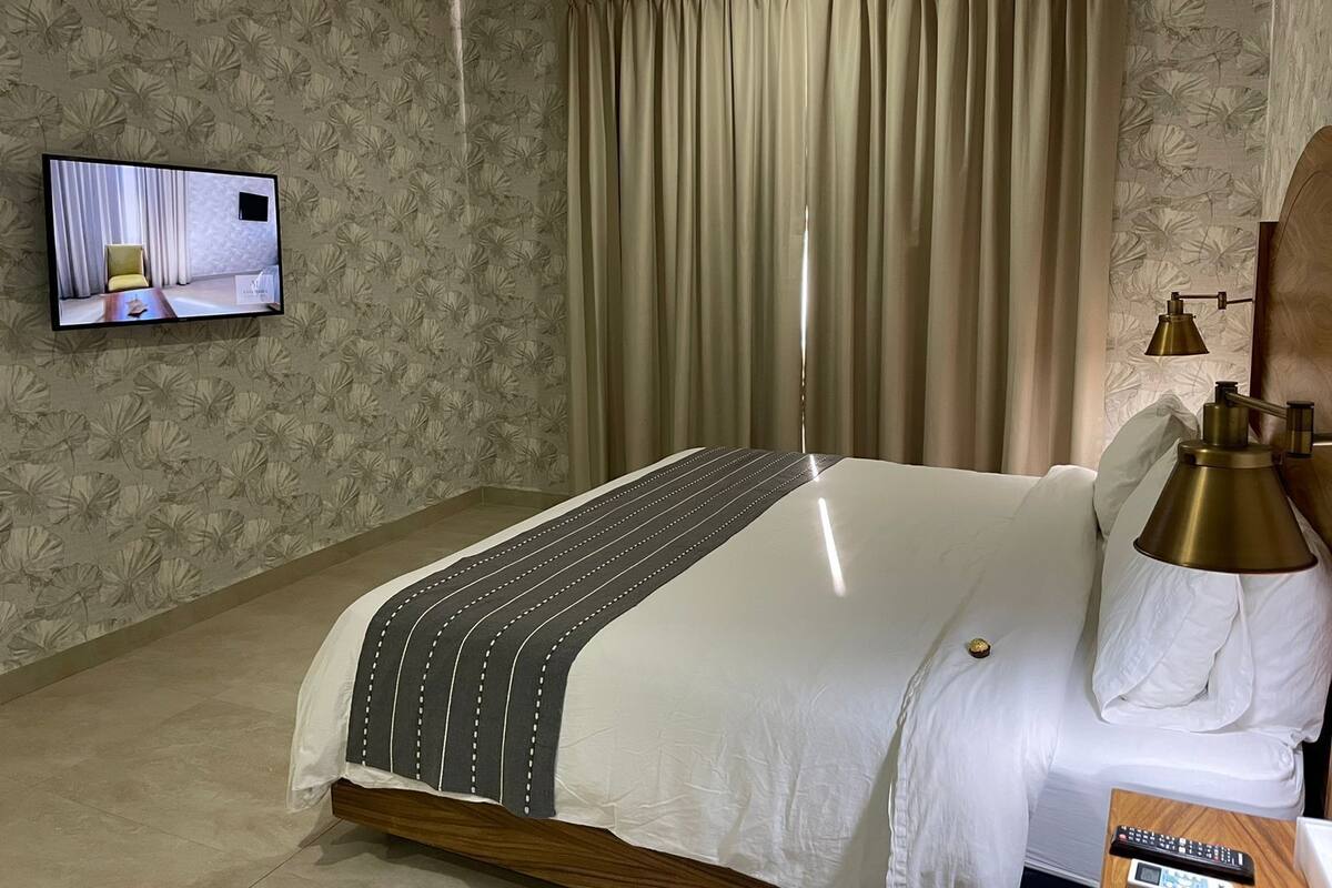 Une chambre d’hôtel avec un lit, des rideaux, un téléviseur fixé au mur et une table de chevet avec une lampe.