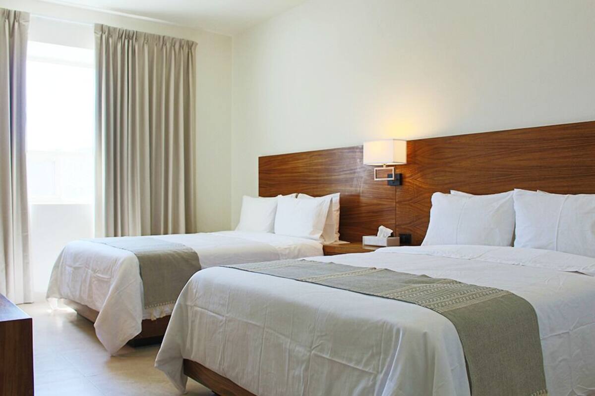 Une chambre d’hôtel avec deux lits, une tête de lit en bois, une lampe de chevet et une fenêtre avec des rideaux.