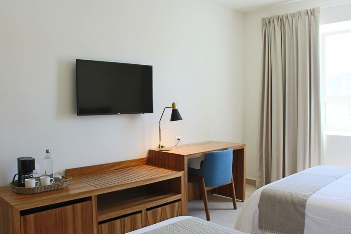 A hotel room with a wooden desk, a chair, a television, a bed, and a window with curtains.