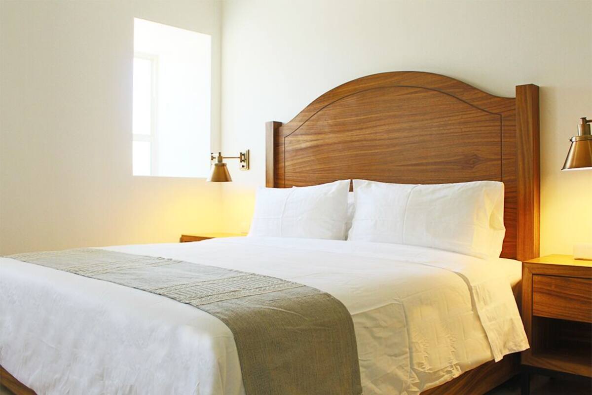 Un lit bien fait, avec une tête de lit en bois, du linge de lit blanc et un plaid de couleur claire. Il y a deux tables de chevet avec des lampes.